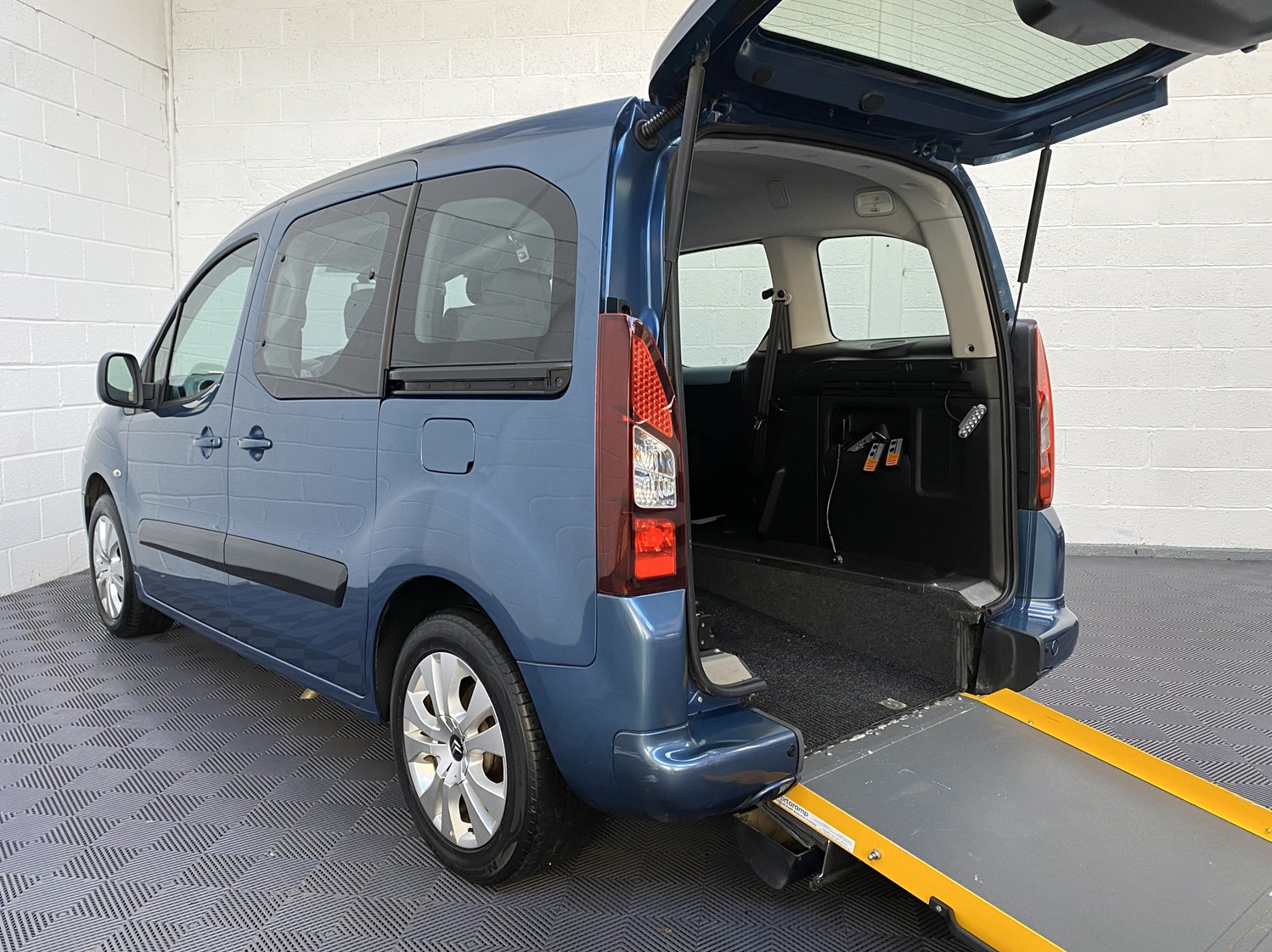 Blue Citroën Berlingo wheelchair accessible vehicle with rear tailgate open and yellow-edged access ramp deployed, showing spacious rear interior, photographed indoors at Michael Harraway Cars NK13 EUW